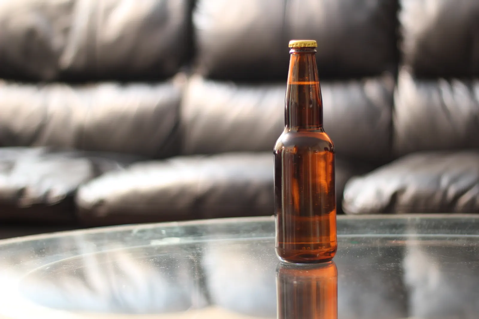 Amber beer bottle on a glass table in front of a blurred black leather sofa