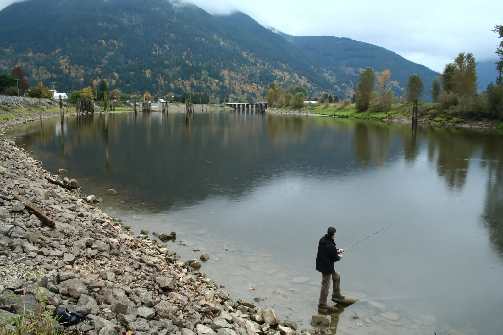 Man fishing with a rod from a rocky shore beside a calm river with forested hills behind