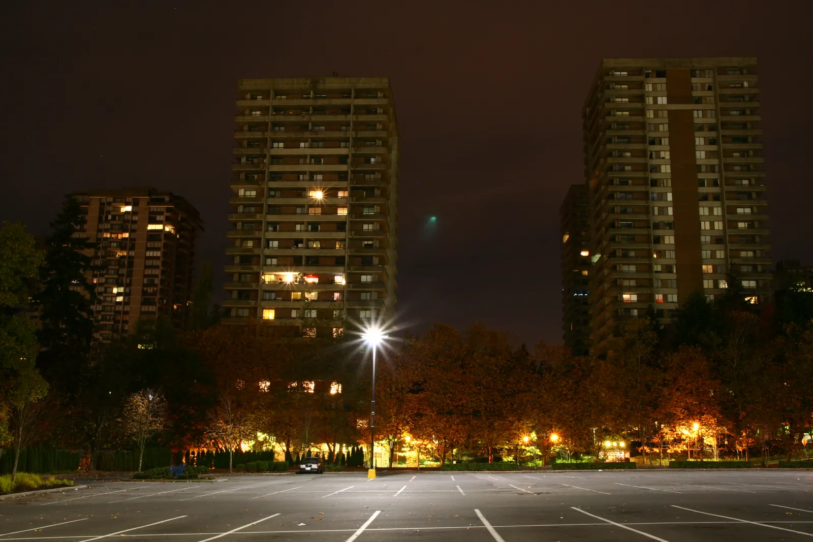 Apartment towers lit at night behind trees and a streetlight above an empty parking lot