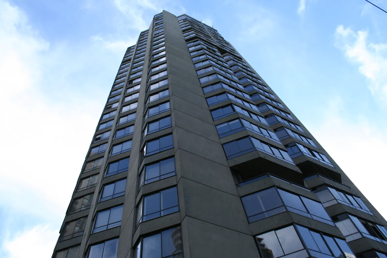 Grey concrete high-rise apartment building with bay windows seen from street level under cloudy sky