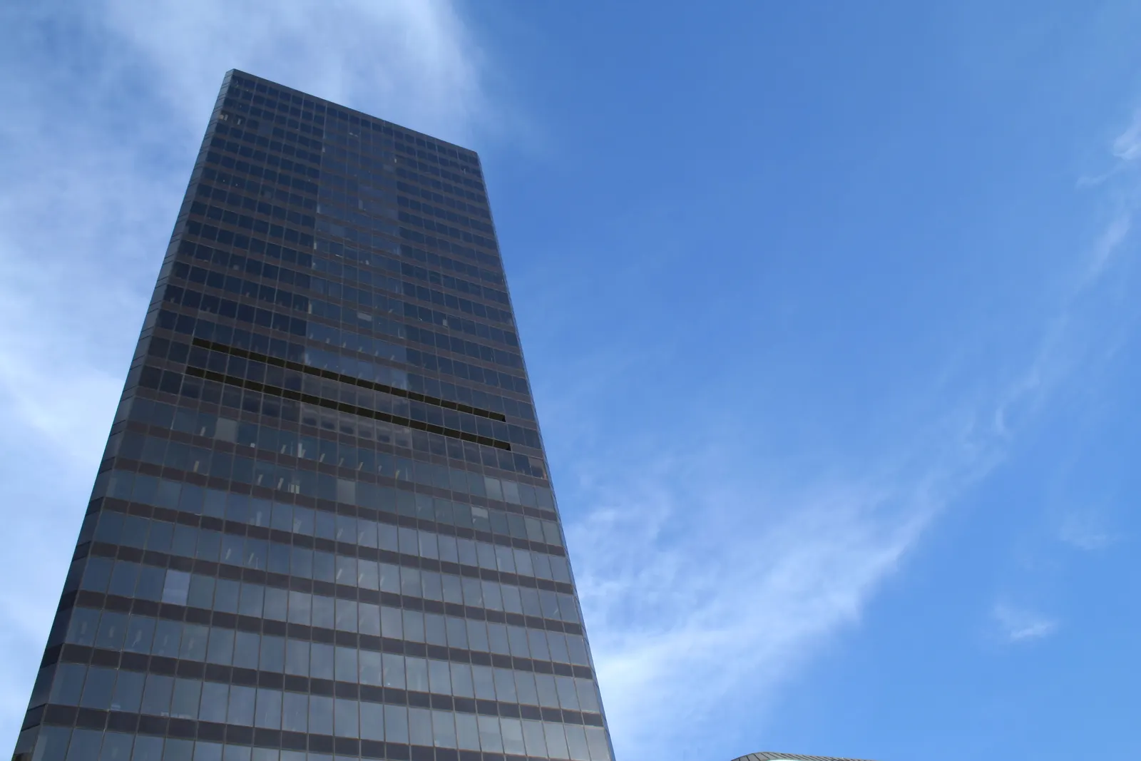 Tall dark glass office skyscraper viewed from below against a blue sky with thin clouds