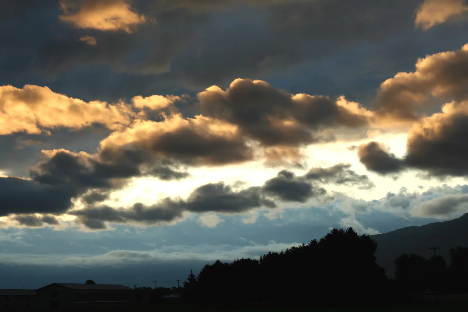 Gold-lit clouds at sunset above a silhouetted tree line and barn