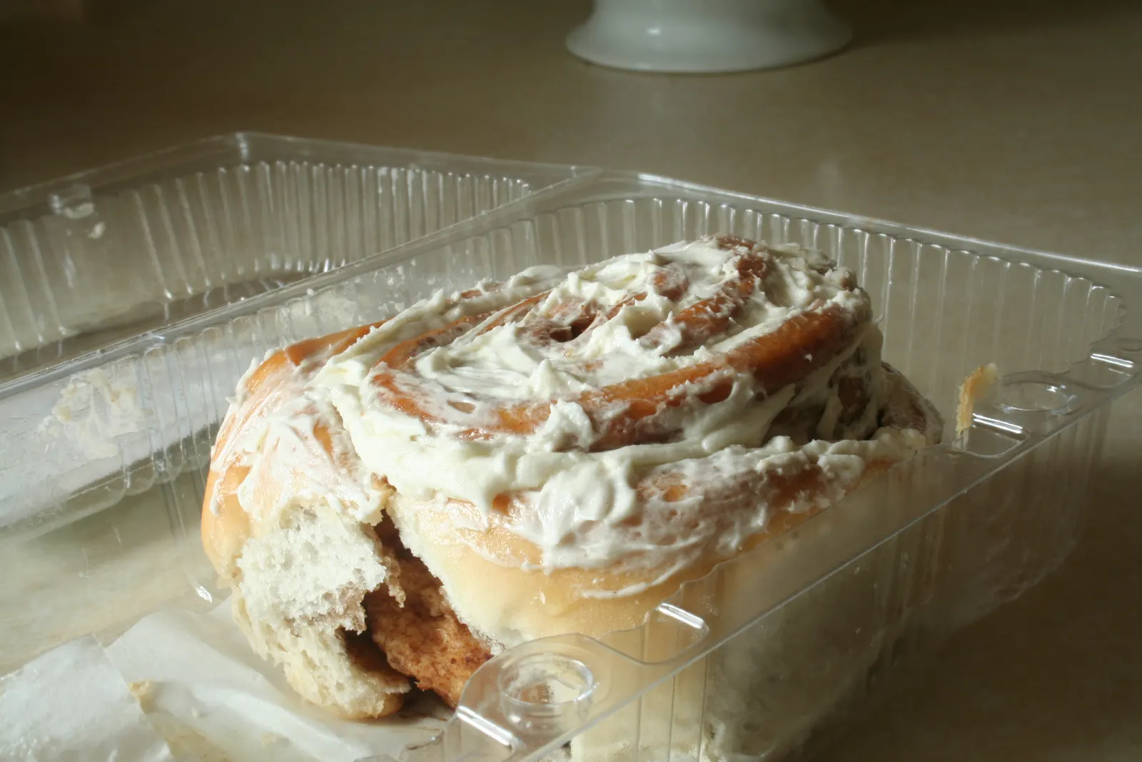 Frosted cinnamon bun in an open clear plastic bakery container on a counter