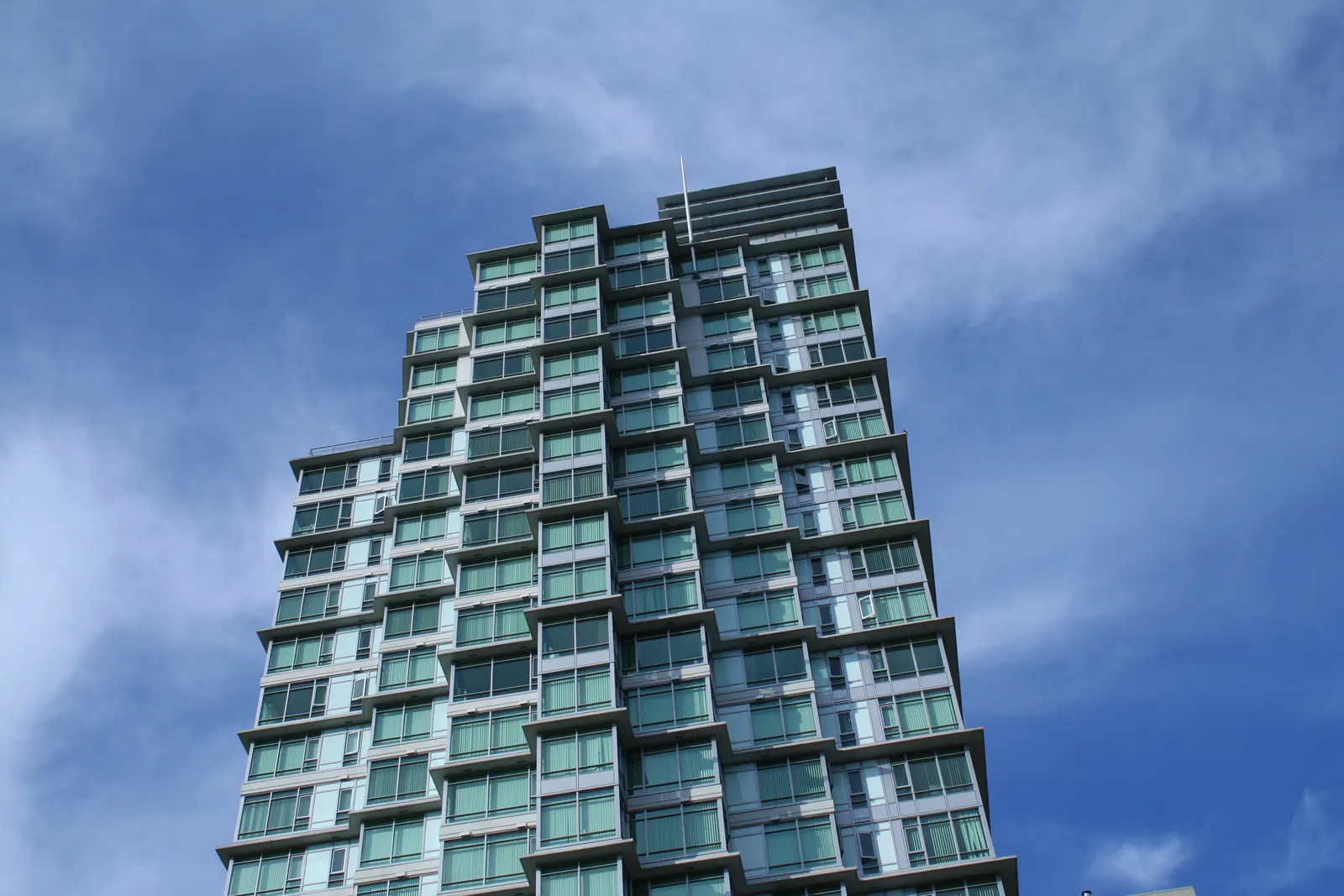 Tall glass residential tower with balconies seen from below against a blue sky with clouds