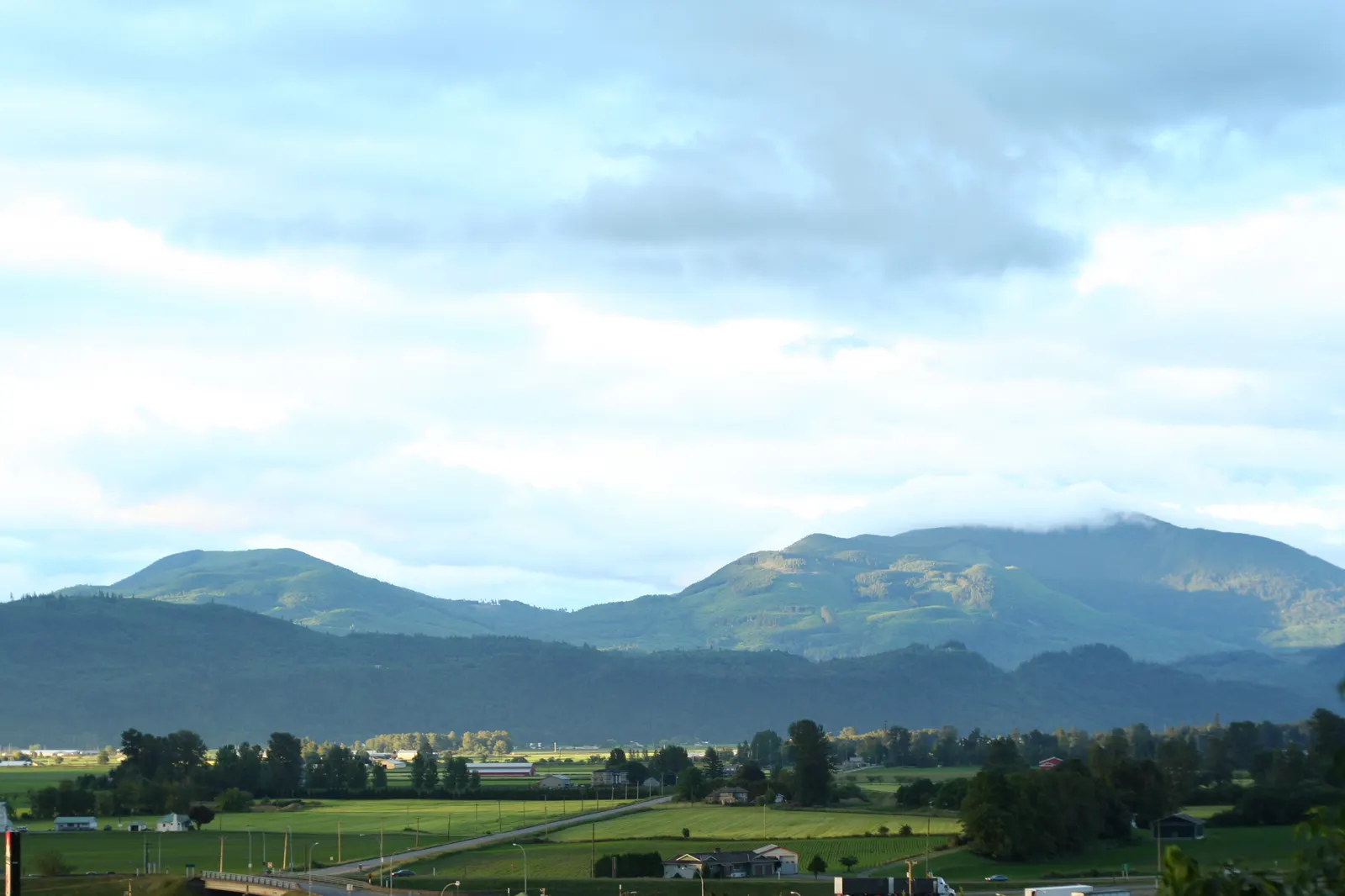 Green valley farmland with barns below forested mountains and a cloudy sky