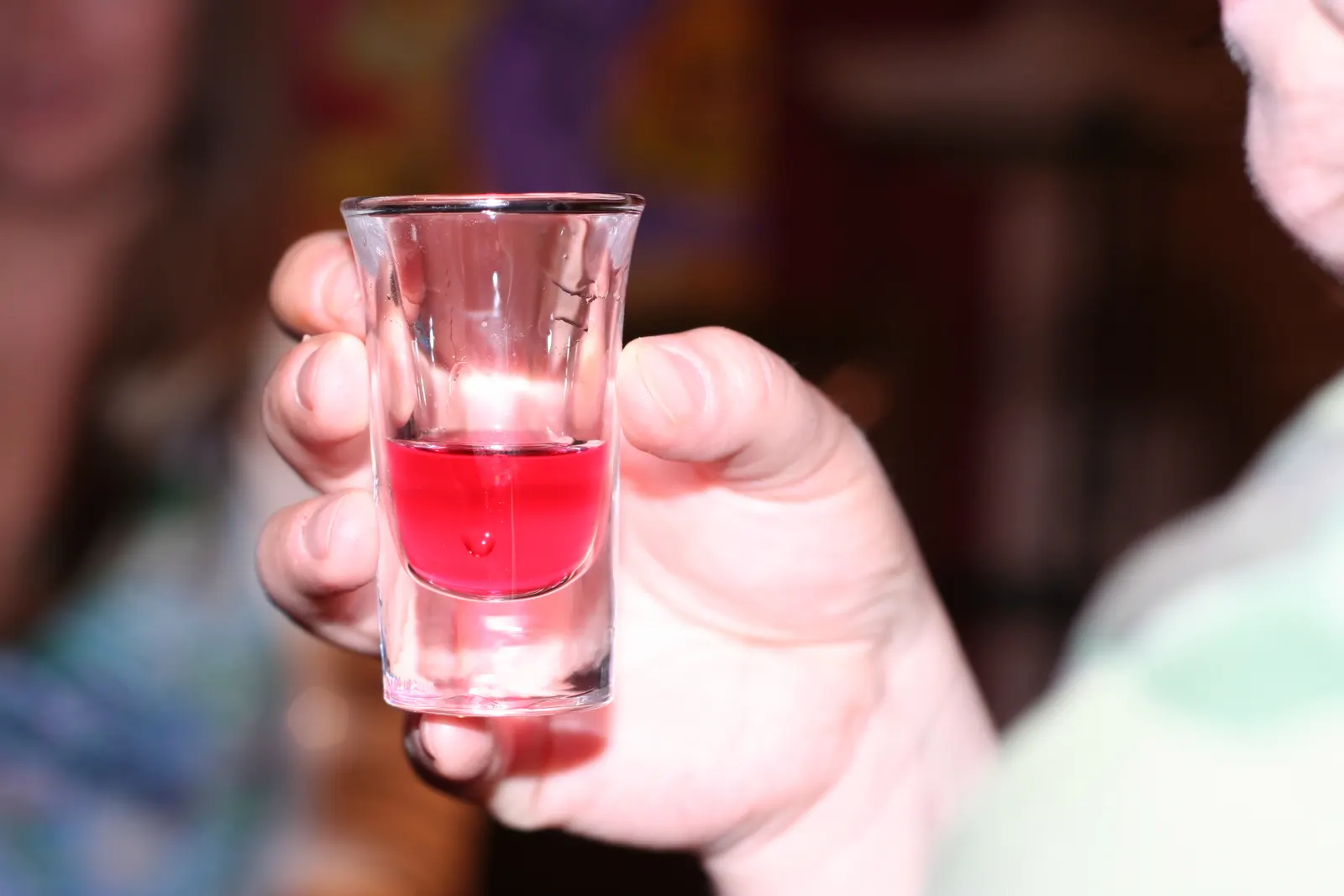 Hand holding a small glass of red liquid toward the camera with blurred people behind