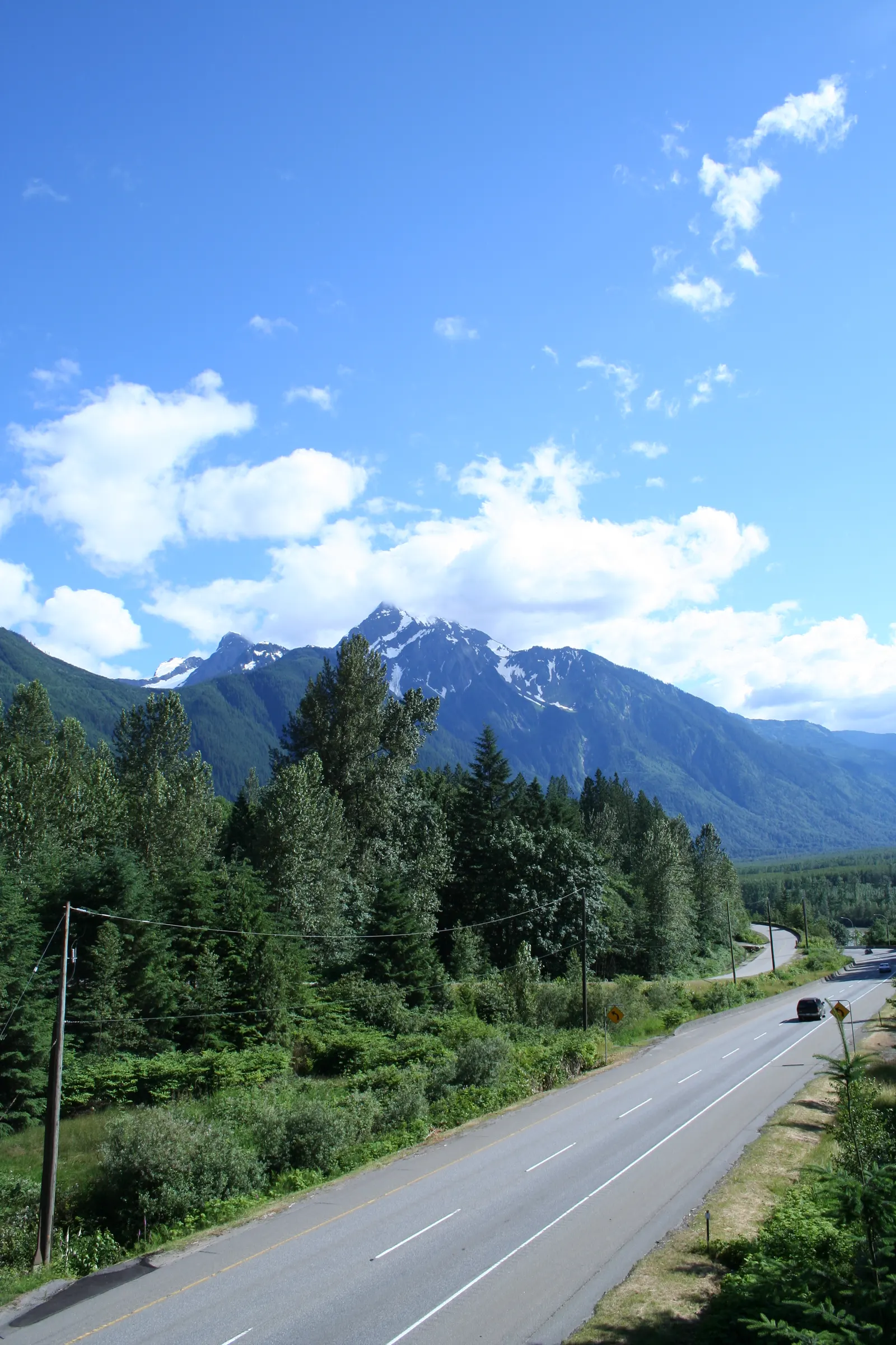 Highway through evergreen forest leading toward snow-capped mountains under blue sky