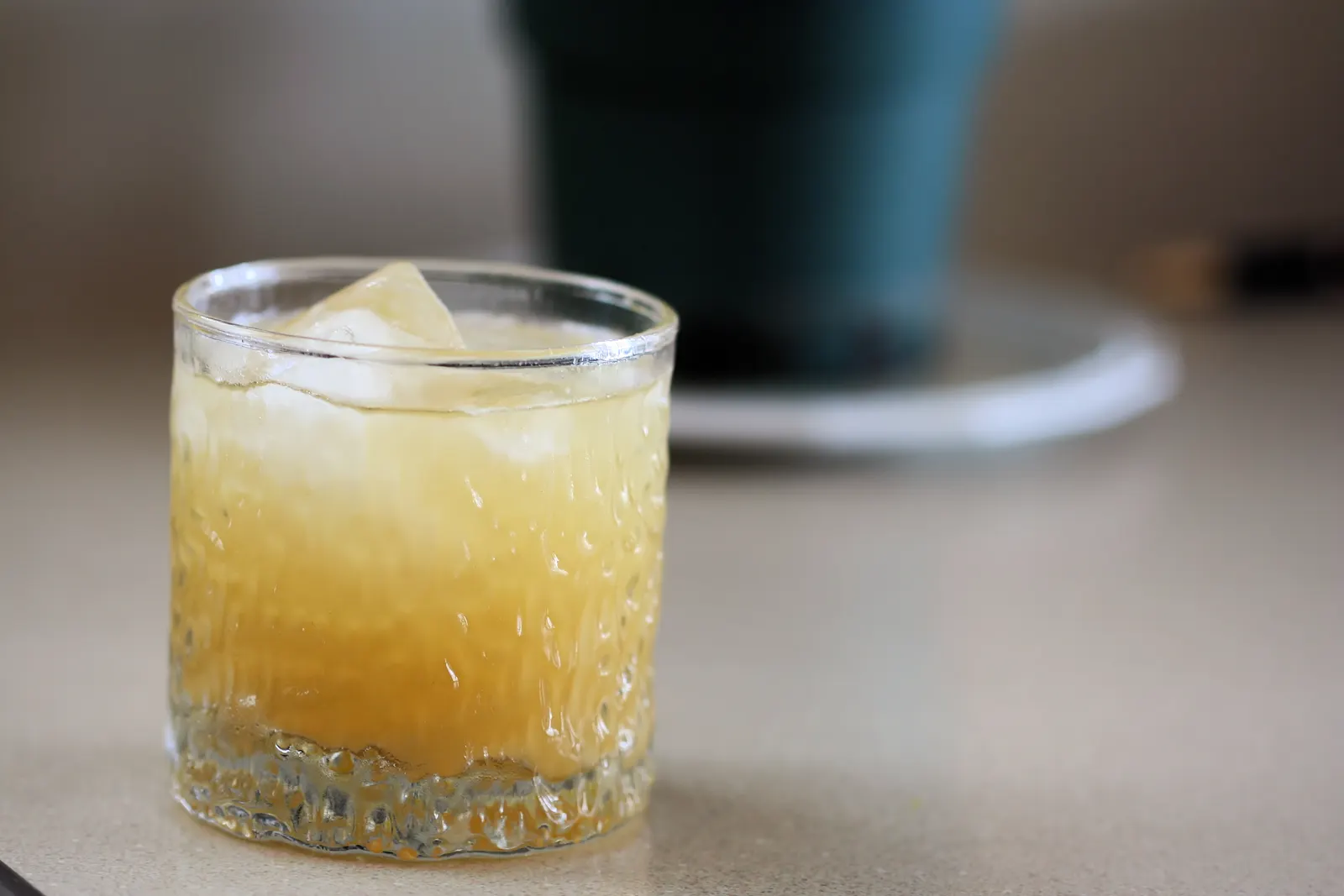 Glass of golden iced drink on a beige countertop with a blurred dark pot behind