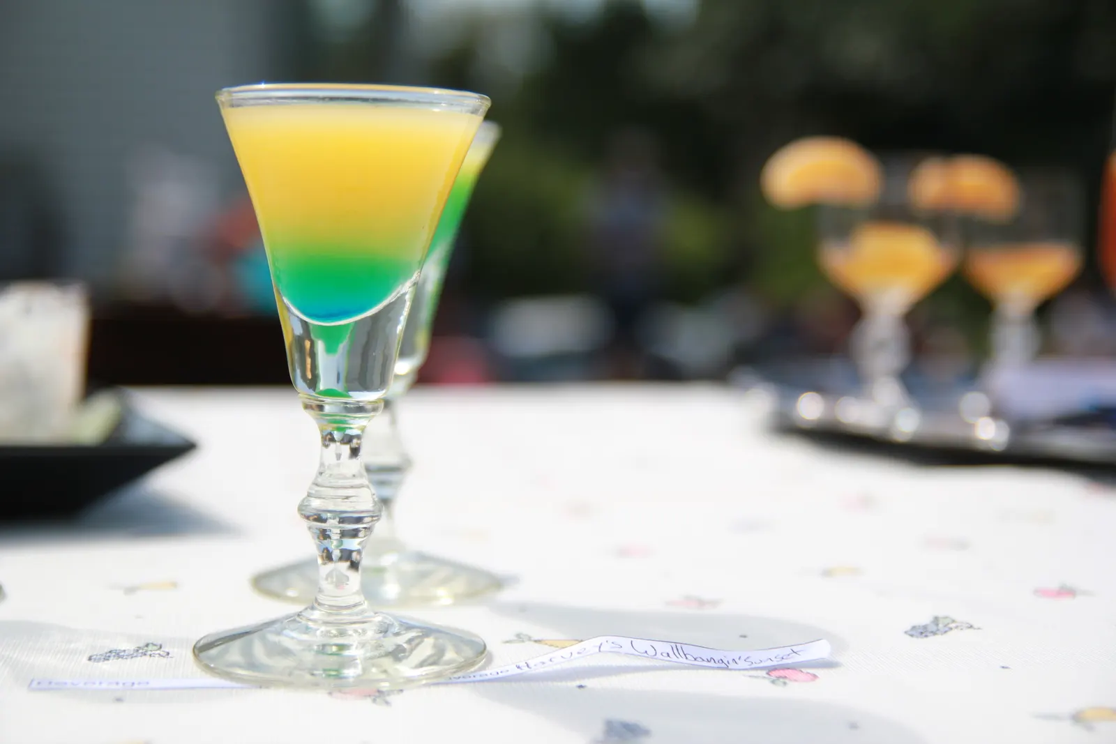 Layered yellow and green cocktail in a stemmed glass on an outdoor table with blurred glasses behind