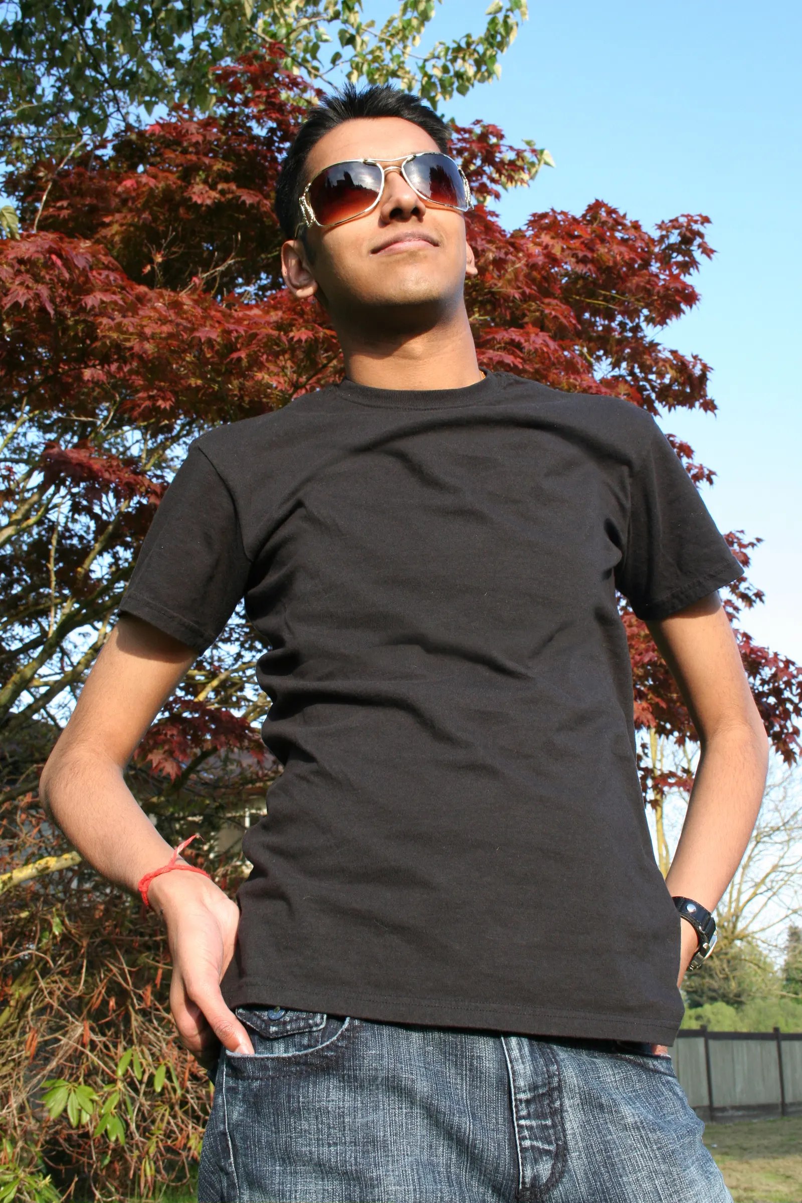Young man in sunglasses, black t-shirt and jeans standing in front of a red maple tree