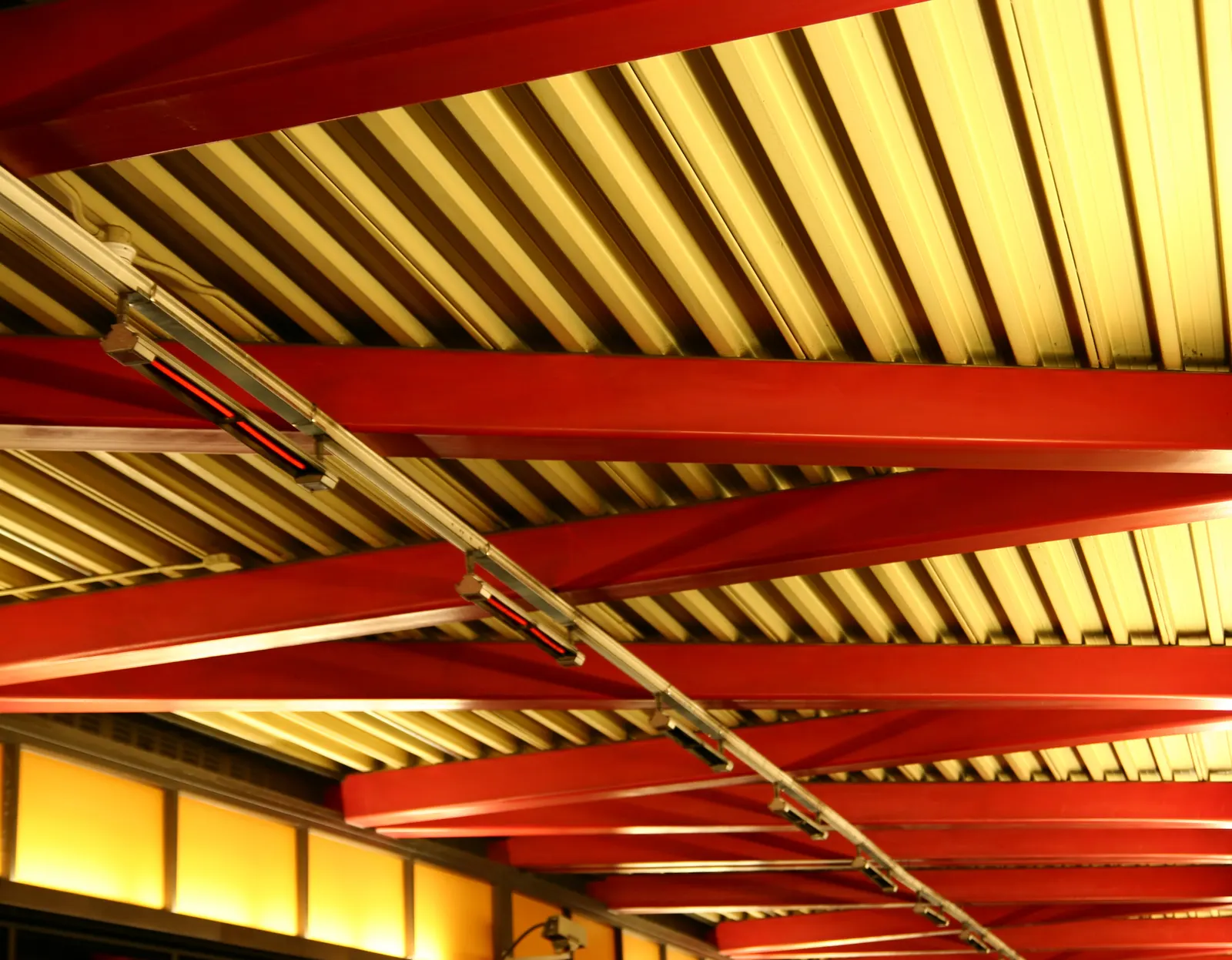 Looking up at red steel beams, ribbed ceiling panels and radiant heaters with amber lights