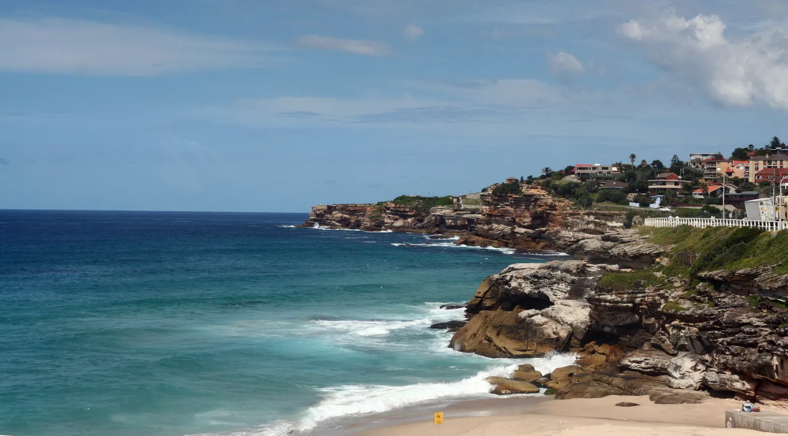 Turquoise ocean waves breaking below sandstone cliffs with clifftop houses under a blue sky