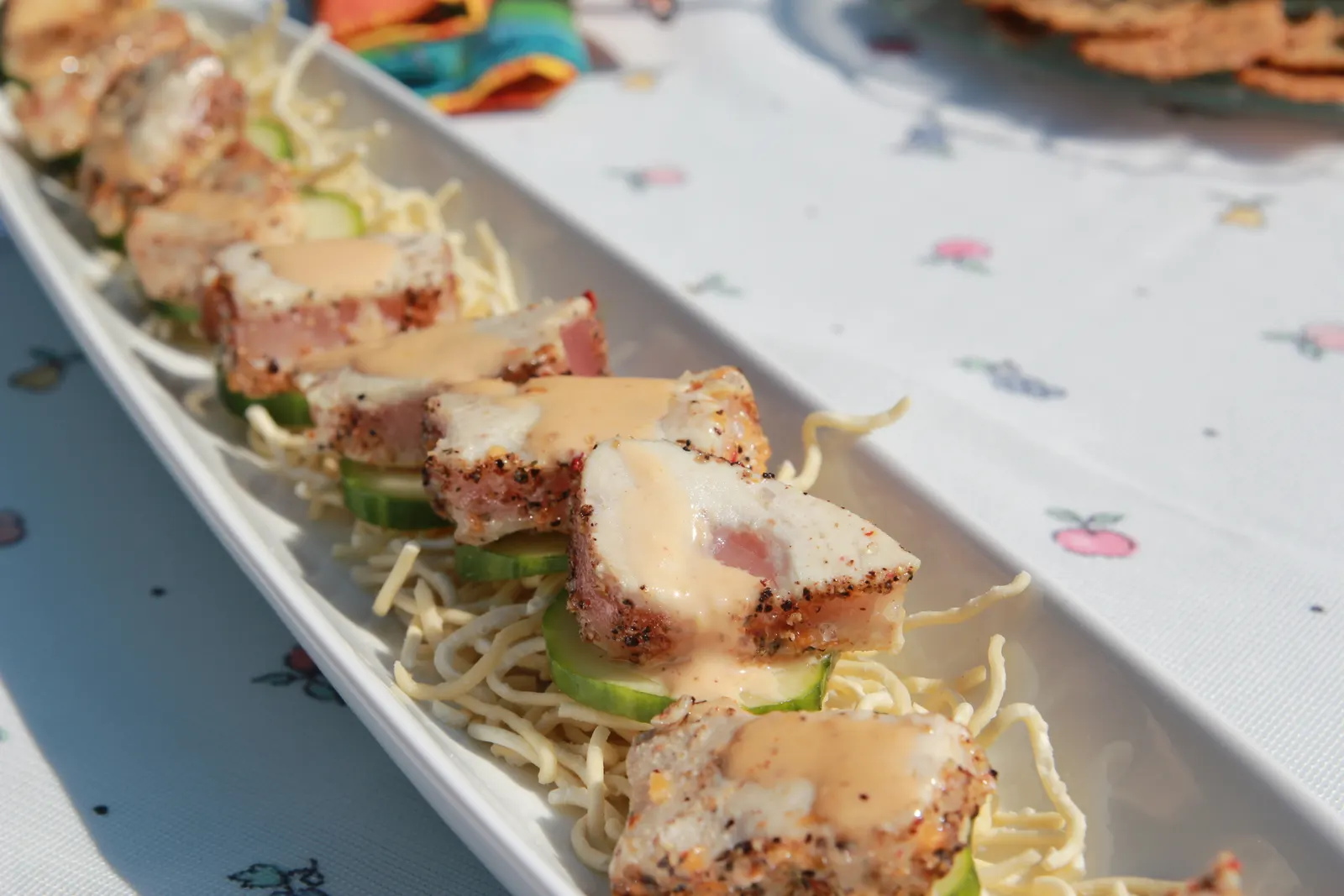 Pepper-crusted seared tuna slices with sauce on cucumber and crispy noodles on a white platter