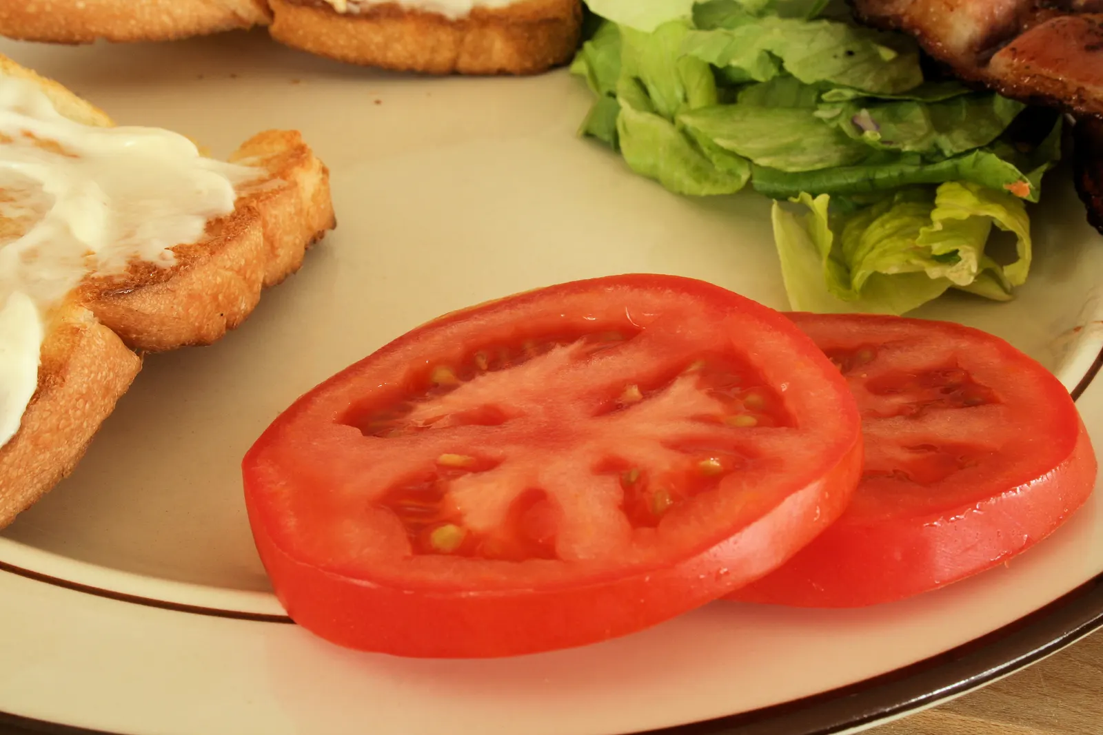Two slices of ripe red tomato on a plate with lettuce, toast and bacon