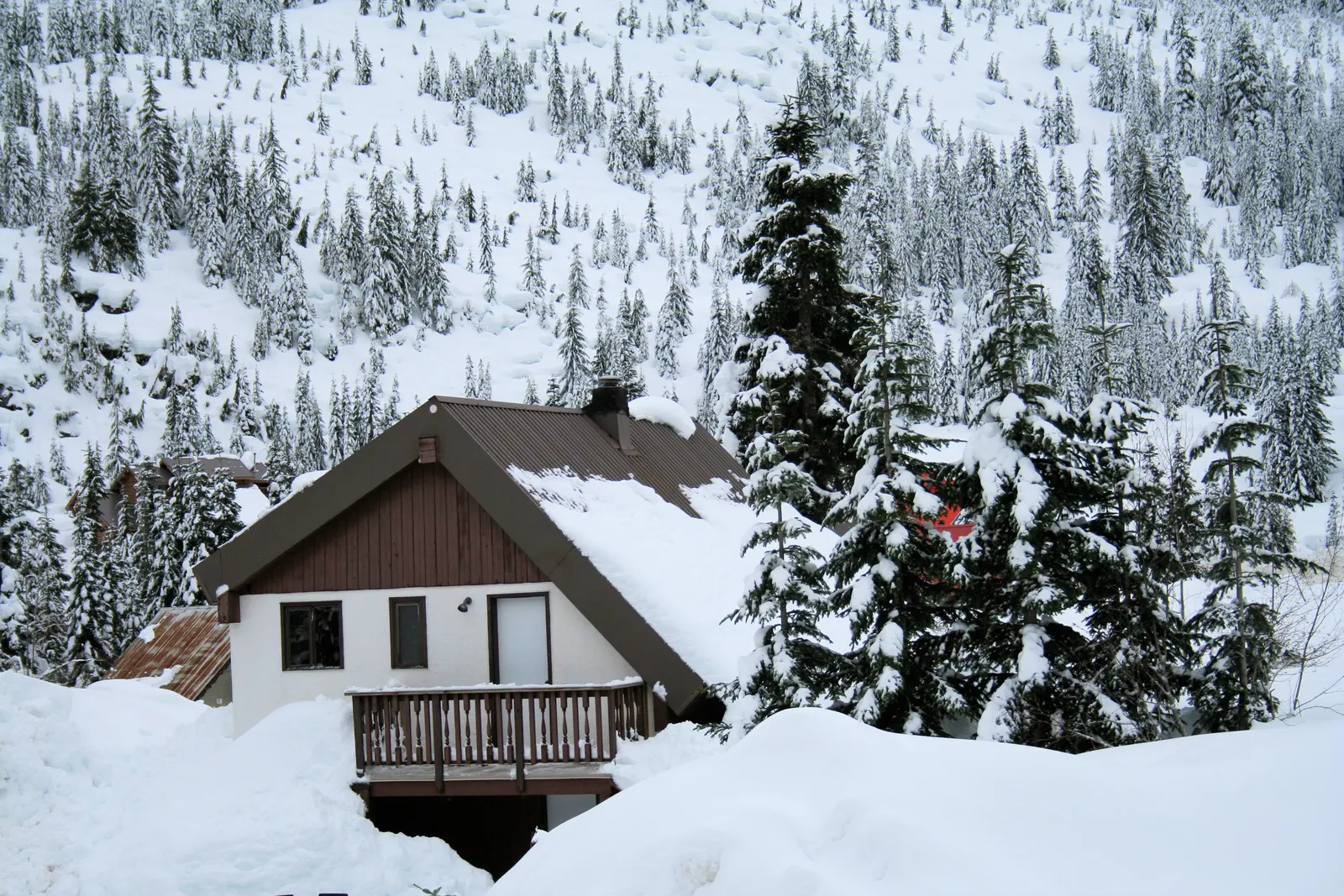 Snow-covered chalet cabin with balcony in deep snow below a hillside of frosted fir trees
