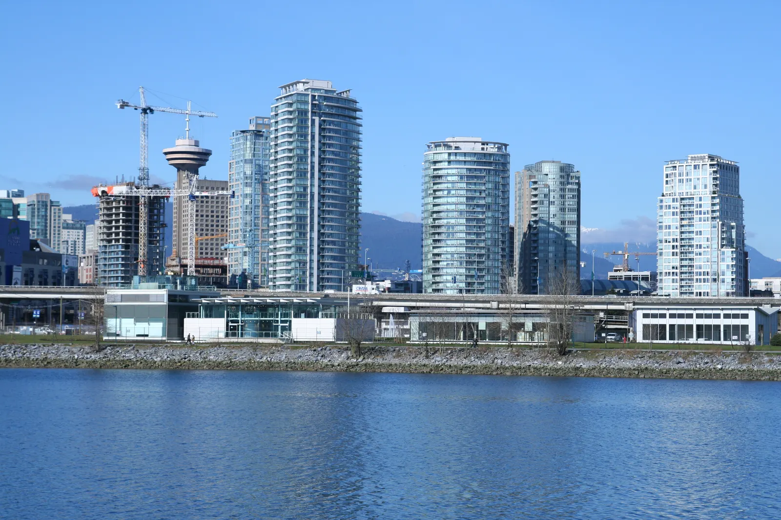 Vancouver downtown glass towers, cranes and mountains beyond a rocky shoreline and calm blue water