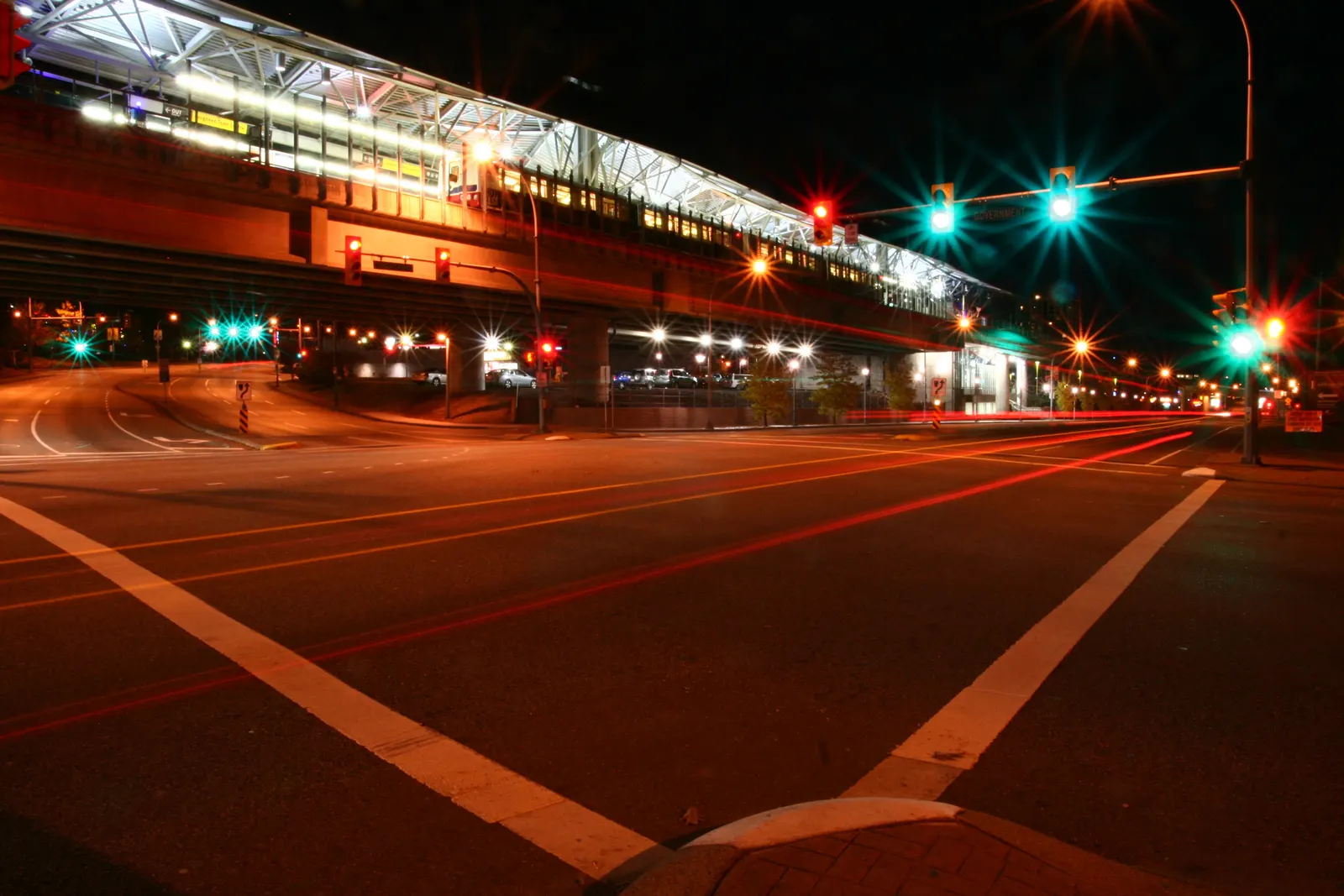 Elevated SkyTrain station at night above an intersection with red light trails and traffic lights