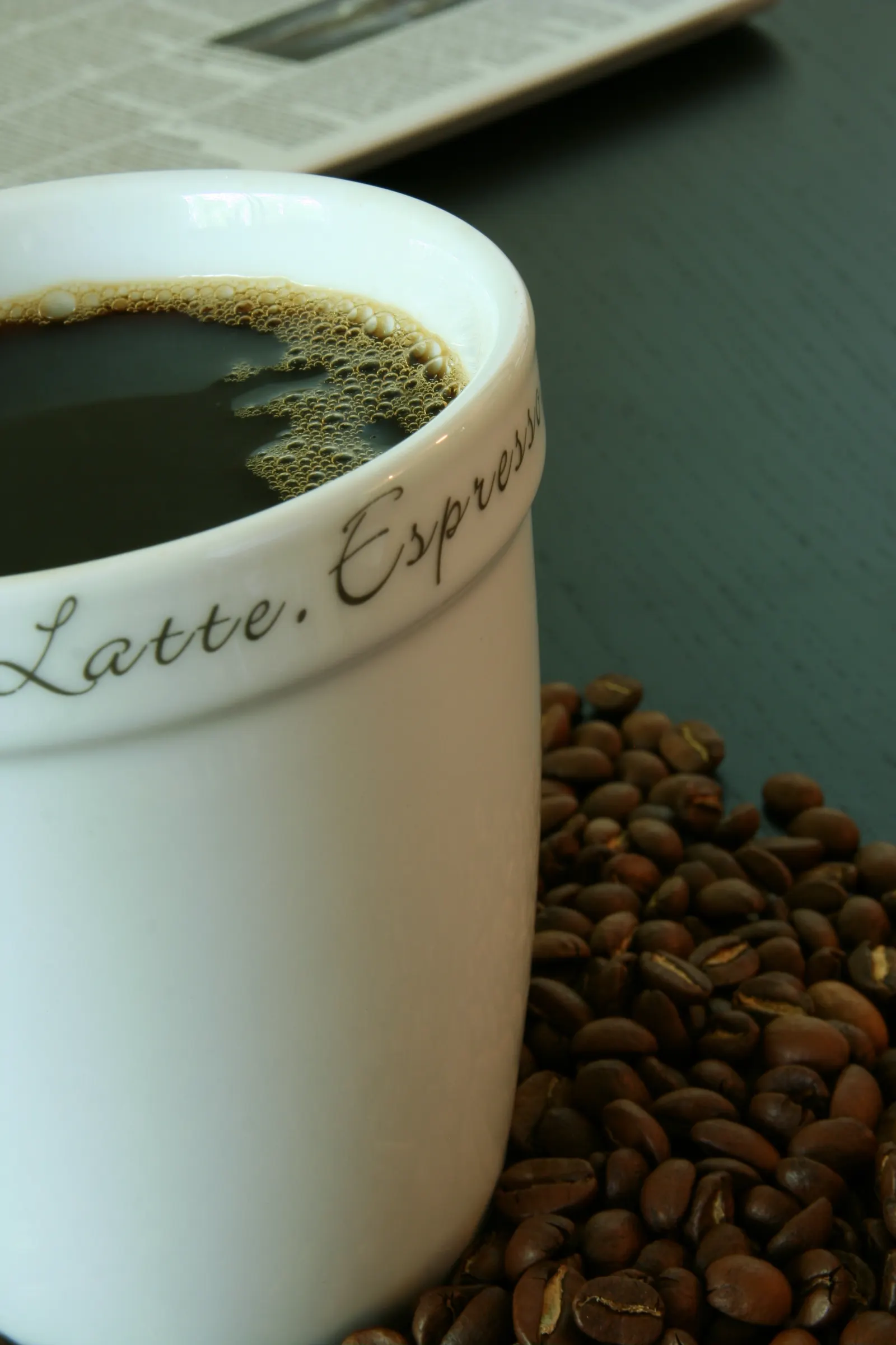 White mug of black coffee with a scattering of roasted coffee beans and a newspaper behind