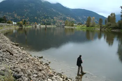 Man fishing with a rod from a rocky shore beside a calm river with forested hills behind