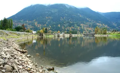 Calm river reflecting a forested autumn mountain, with a rocky shore and bridge in the distance
