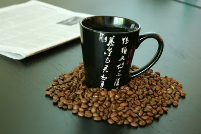 Black ceramic mug of coffee with white calligraphy sitting on scattered roasted coffee beans