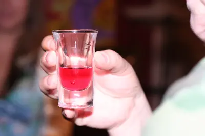 Hand holding a small glass of red liquid toward the camera with blurred people behind