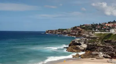 Turquoise ocean waves breaking below sandstone cliffs with clifftop houses under a blue sky