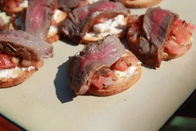 Sliced rare steak on toasted rounds with soft cheese and diced tomato on a cream plate