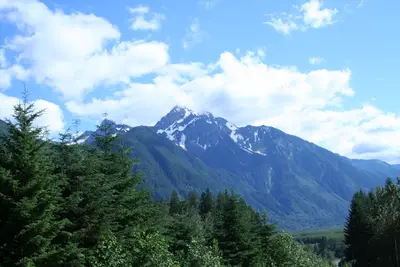 Snow-capped mountain peak above evergreen forest under a blue sky with white clouds