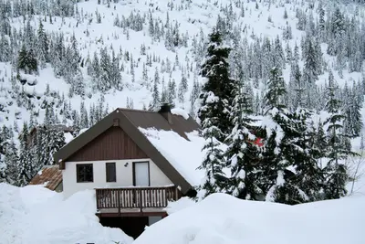 Snow-covered chalet cabin with balcony in deep snow below a hillside of frosted fir trees