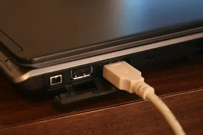 Beige USB cable plugged into the side of a black laptop on a wooden desk