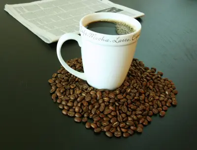 White coffee mug sitting on a pile of roasted coffee beans beside a folded newspaper