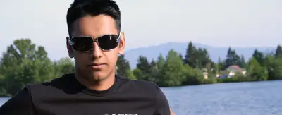 Young man wearing sunglasses and a black t-shirt in front of a lake and trees